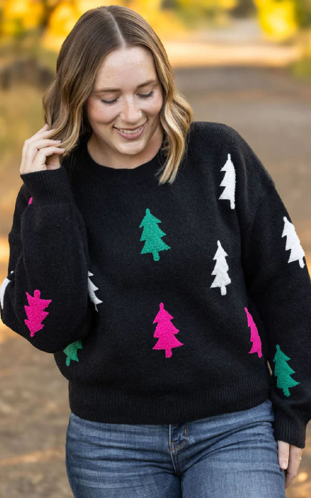 Woman modeling a whimsical holiday sweater that is black and adorned with white, green and pink christmas trees.