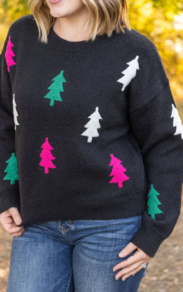 Woman modeling a whimsical holiday sweater that is black and adorned with white, green and pink christmas trees.