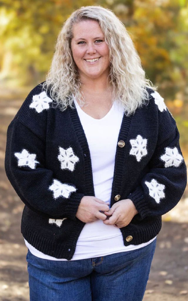 Woman wearing a whimsical holiday sweater that is black with white applique snowflakes. 