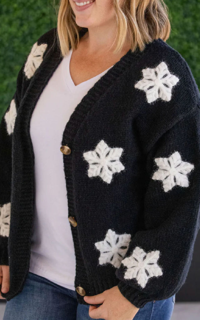 Woman wearing a whimsical holiday sweater that is black with white applique snowflakes. 